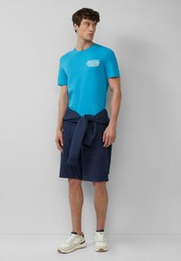 Turquoise T-shirt met korte mouwen met een witte graphic, gecombineerd met marineblauwe shorts die om de taille zijn geknoopt. Witte sportieve schoenen maken de outfit compleet.
