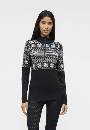 Mujer que lleva una camiseta de manga larga con patrón en blanco y negro con media cremallera y leggins negros a juego con el mismo patrón, de pie frente a un fondo liso.