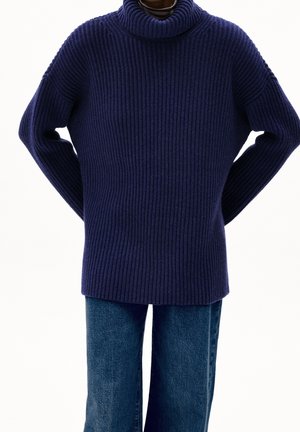 Person wearing a chunky navy blue ribbed turtleneck sweater and straight-cut blue denim jeans, hands behind back.