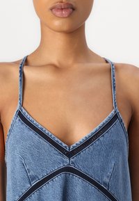 Denim top med tynne stropper, V-utringning, og kontrasterende marineblå kant rundt bysten, med et lys blått teksturert stoff.