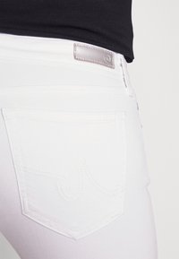 Jeans de mezclilla blanca con un bolsillo trasero que presenta un patrón de costura decorativo y un parche de cuero plateado en la cintura.