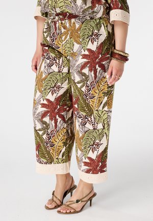 Femme portant un pantalon court imprimé de feuilles tropicales vertes, rouges et jaunes, détails de poignets beiges, bracelets dorés et sandales à talons marron avec des lanières.