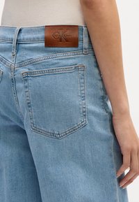 Šviesiai mėlynos džinsinės kelnės pasižymi lygia tekstūra, klasikinio keturių kišenių dizainu ir rudu odiniu Calvin Klein logotipo ženkleliu ant juosmens.