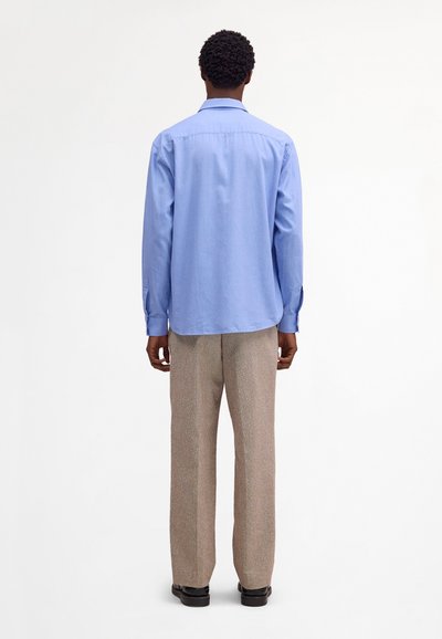 Uomo visto da dietro che indossa una camicia a maniche lunghe azzurro chiaro, pantaloni beige e scarpe nere, in piedi su uno sfondo bianco uniforme.