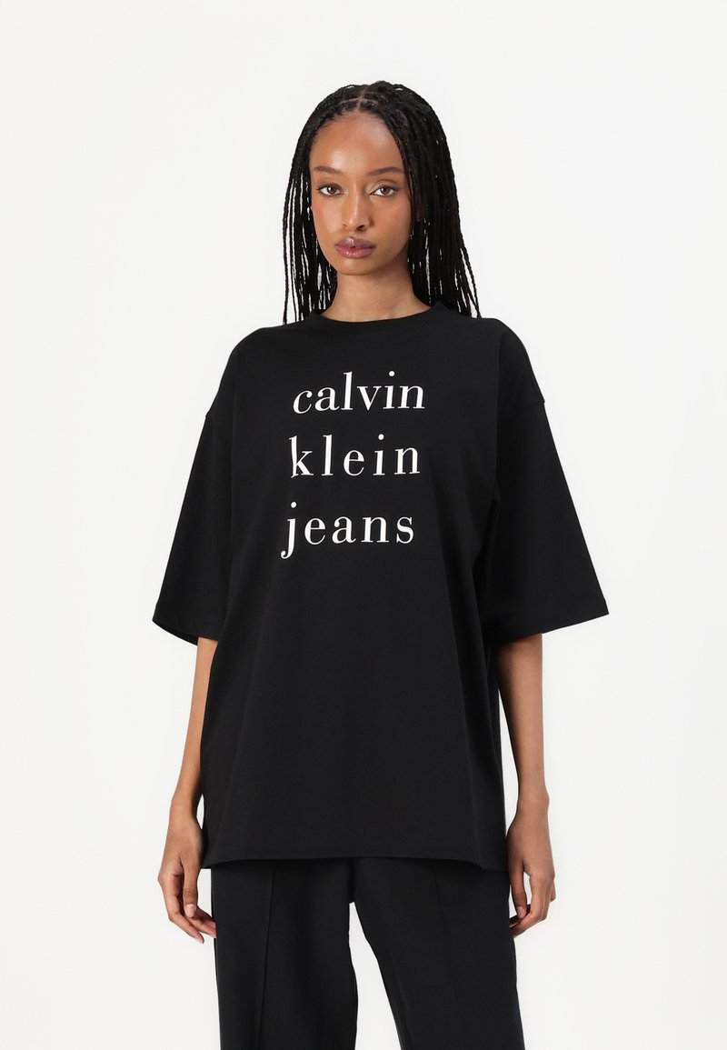 Černé oversized tričko vyrobené z bavlny, s bílým textem, který zní "calvin klein jeans" uspořádaným ve sloupcovém formátu. Krátké rukávy.