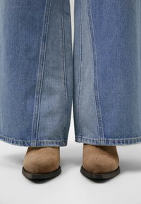 Botas de ante marrón hasta el tobillo con punteras redondas, combinadas con jeans de mezclilla azul claro de pierna ancha con costuras en contraste y dobladillos deshilachados.