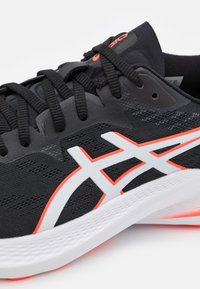 Zapatilla deportiva negra con parte superior de malla y superposición blanca que presenta un logo, acentuada con detalles en naranja. Suela texturizada con espuma blanca.