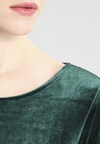 Zielona aksamitna bluzka w odcieniu emerald z gładką fakturą i szerokim dekoltem. Widoczne detale przeszyć wzdłuż szwu na ramieniu.