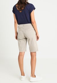 Kvinna som bär en mörkblå kortärmad topp med korsade detaljer i ryggen, beige knälånga shorts och vita slip-on sneakers, stående vänd bort.