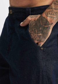Tummat farkut, joissa on etutasku, johon käsi on osittain työnnetty. Näkyvissä ompeleet ja metallinen nappi, tatuoitu käsi.