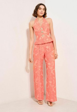 Frau trägt ein korallenfarbenes, geblümtes Neckholder-Top mit drei dekorativen Knöpfen und passende Hosen mit weitem Bein, steht vor einer hellen Wand.