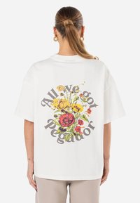 Hvit oversized t-skjorte med et fargerikt blomsterprint og teksten "All We got Pegador" i en sirkulær formasjon på baksiden.