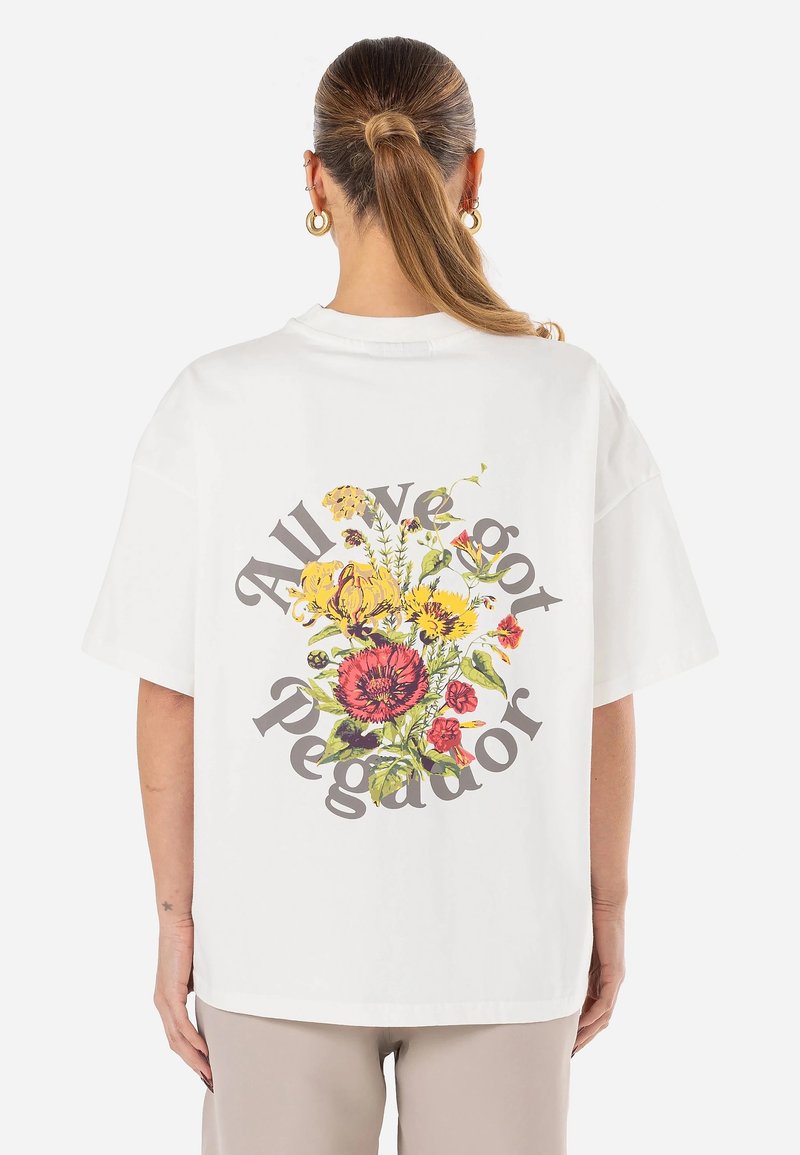Hvit oversized t-skjorte med et fargerikt blomsterprint og teksten "All We got Pegador" i en sirkulær formasjon på baksiden.