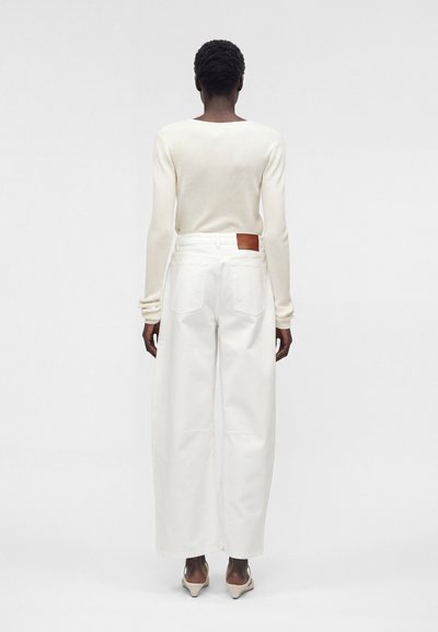 Άτομο στέκεται με την πλάτη, φοράει μια κρεμ ριπ μπλούζα με μακριά μανίκια, λευκά φαρδιά παντελόνια με τσέπες πίσω και μπεζ παπούτσια με τακούνι.