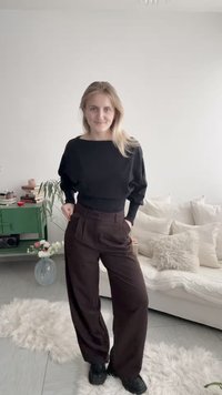 Donna con maglione nero e pantaloni larghi scuri in piedi in un soggiorno luminoso con divano bianco, mobile verde e fiori in un vaso di vetro.