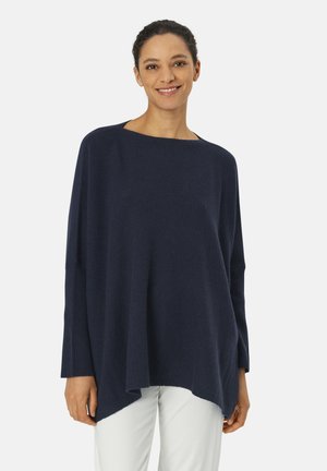 Maglione oversize blu navy con una ampia scollatura a barca, polsini a coste e un orlo asimmetrico, abbinato a pantaloni di colore chiaro.