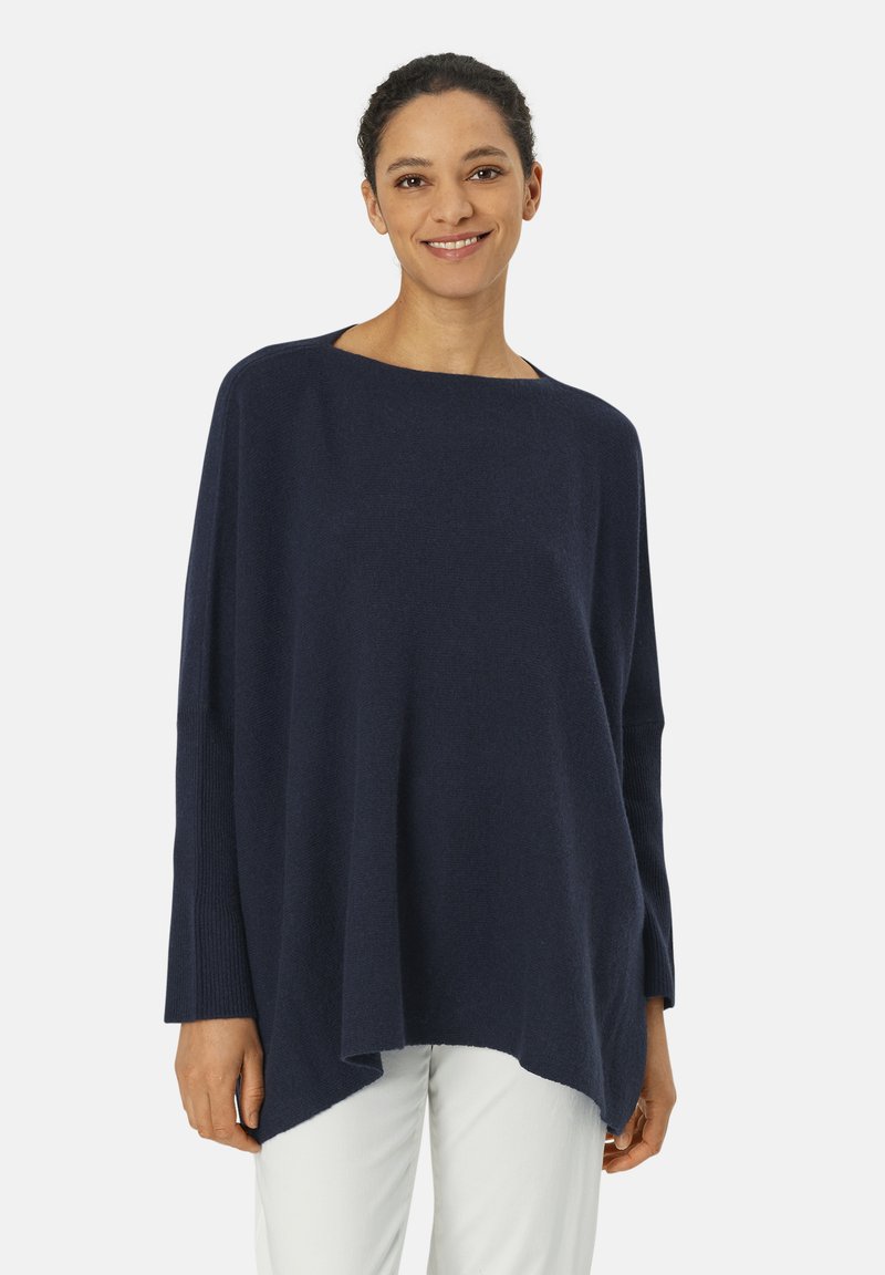 Marinefarbener, übergroßer Strickpullover mit weitem Bootsausschnitt, gerippten Ärmelbündchen und asymmetrischem Saum, kombiniert mit hellen Hosen.