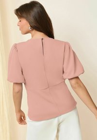 Blusa rosa chiaro con maniche a sbuffo e dettaglio a chiave sul retro, realizzata in tessuto morbido. Abbinata a pantaloni bianchi con texture morbida.