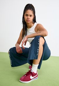 Mėlynos kelnės su baltu Nike logotipu, derinamos su bordo spalvos Nike sportbačiais ir balta maike be rankovių. Supinto plaukai ir paprastas kaklo papuošalas.