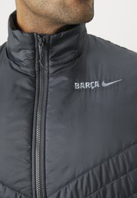 Black puffer jacket with high collar, zip closure, and quilted texture. Features silver "BARÇA" and Nike logos on the chest.