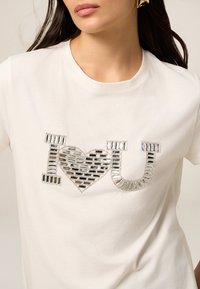 Camiseta blanca de algodón con un gráfico de lentejuelas plateadas "I ❤️ U" en el frontal, con detalles texturizados y acentos reflectantes.