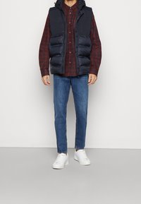 Laivastonsininen toppaliivi hupulla punaisen ja mustan ruudullisen paidan päällä, yhdistetty sinisiin farkkuihin ja valkoisiin lenkkareihin. Ominaisuuksiin kuuluu vetoketjullinen sulkeminen ja taskut.