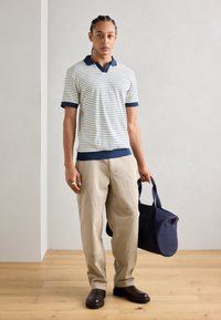 Polo rayé avec un col marine, pantalon beige et chaussures marron foncé ; tenant un sac marine. Texture douce ; coupe décontractée.