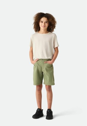 Barn med krøllet hår iført beige kortærmet skjorte, olivengrønne shorts og sorte sneakers, står med hænderne i lommerne.