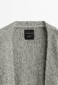 Pull en maille texturée grise avec col plié et étiquette noire indiquant "Massimo Dutti Studio" à l'encolure.