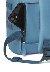 Zaino in tessuto blu chiaro con texture, caratterizzato da un telefono parzialmente visibile in una tasca con zip, con una tracolla scura sul lato.