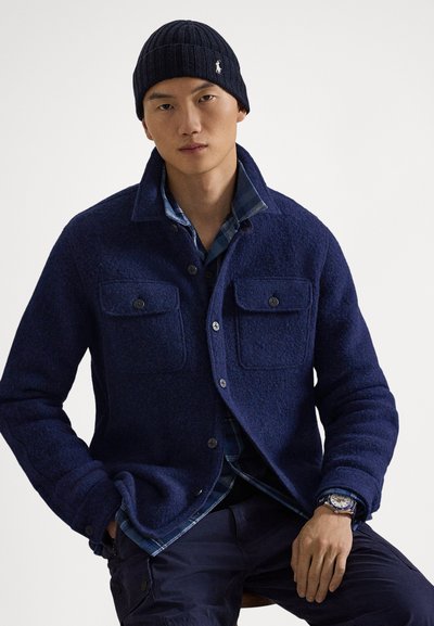 Marineblaue Jacke aus Wollmischung mit zwei Brusttaschen, vorne mit Knöpfen zu schließen und mit gerippten Bündchen. Darüber ein blau kariertes Hemd und dazu eine schwarze Strickmütze.