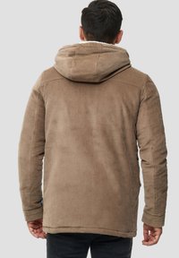 Mand med kort, mørkt hår iført en tan corduroy hættejakke og sorte bukser, vendende væk fra en ensfarvet grå baggrund.