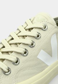 Sneaker in tela beige con logo bianco, occhielli argento e lacci piatti. Presenta una superficie testurizzata e suola in gomma.