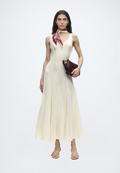 Γυναίκα φοράει αμάνικο εκρού πλεκτό φόρεμα, μπορντό κασκόλ με σχέδια στο λαιμό, κρατά σκούρο καφέ τσαντάκι και άσπρα σανδάλια.