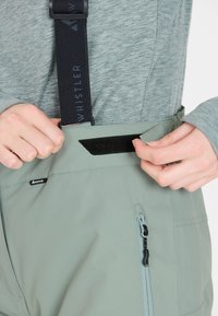 Helle grüne wasserdichte Latzhose mit verstellbaren schwarzen Trägern und einem Klettverschluss an der Taille, ausgestattet mit einer seitlichen Reißverschlusstasche.