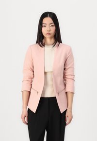 Light pink blazer with a fitted design, round neckline, and 3/4 length sleeves. Features two front zippers and a smooth texture. Paired with a cream ribbed top.