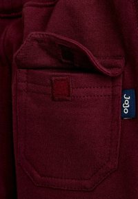 Vinrødt stof med en tekstureret overflade har en rektangulær baglomme og en blå etiket med hvid tekst. Syningerne er pænt udført.