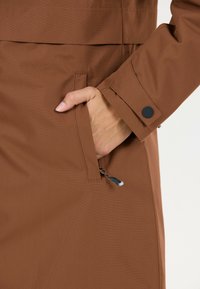 Bruine waterdichte stoffen jas met een zijzak, zichtbaar hand dat in de zak steekt, met een rits en knoopdetail op de mouw.