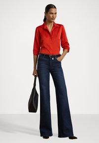Camisa vermelha de botão com mangas roladas, combinada com jeans flare azul escuro. Acessorizada com uma mala tote preta, de textura suave.