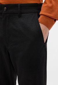 Čierne bavlnené nohavice s hladkou textúrou, so prednou vreckom, zapínaním na gombík a kontrastným oranžovým rukávom z blízkej košele.