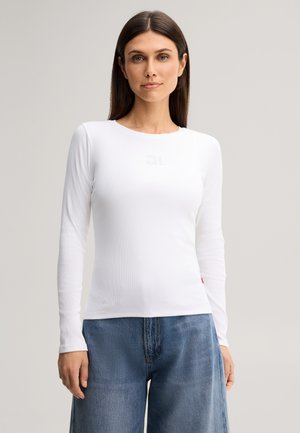 Frau mit glatten dunklen Haaren, die ein eng anliegendes weißes langärmliges Oberteil und weite blaue Jeans trägt, steht vor einem schlichten Hintergrund.