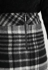 Schwarzes Fleece-Oberteil mit einem grauen Karorock. Verfügt über einen silbernen Reißverschluss, kontrastierende Texturen und eine Mischung aus schwarz-weißen Mustern.