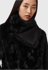 Cappotto di pelliccia nera con colletto in raso nero liscio e dettaglio triangolare sul retro. Presenta una texture morbida e lunga della pelliccia e una chiusura con bottoni.