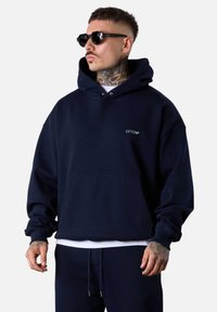 Pull à capuche bleu marine avec une poche avant et un détail de logo blanc sur la poitrine. Associé à un pantalon bleu marine assorti. La texture semble douce.