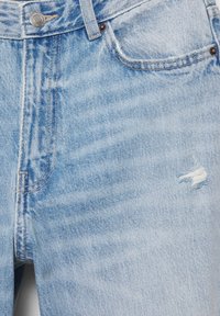 Ljusblå jeans i denim, närbild som visar framficka, metallknapp, bältesögla och en liten sliten reva på höger lår.