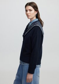 Maglione a maglia blu navy con cappuccio a motivo, polsini e orlo a coste, indossato sopra una camicia colletto azzurro; vestibilità rilassata.