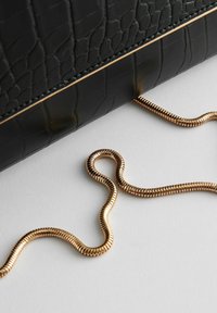 Bolso de mano de cuero negro con relieve de cocodrilo y cadena dorada, curvado sobre una superficie blanca. Presenta un acabado texturizado y un diseño elegante.