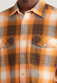 Camicia in flanella a quadri arancione e marrone con due tasche sul petto, chiusura a bottoni e una texture morbida. Presenta un colletto a punta e motivi a contrasto.