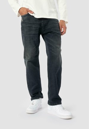 Man in donkere vervaagde jeans met lichte slijtage en witte sneakers, staande met één hand in de zak tegen een effen achtergrond.