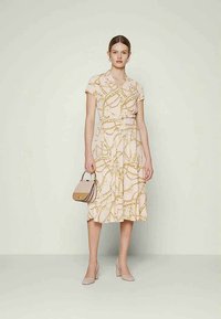 Mulher com um vestido midi cor de rosa pálido com estampado de correntes douradas, segura uma bolsa bege e usa sapatos de salto bloco combinando, de pé contra uma parede clara.